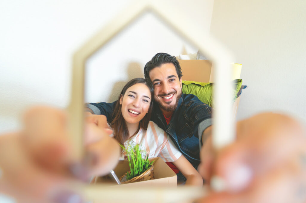 A young Hispanic couple smiling as they hold a small house shape in celebration of owning a new home with help from down payment assistance programs.