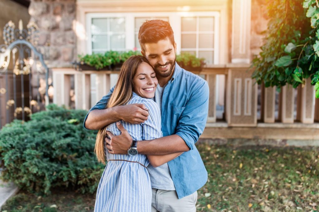 a young couple embracing outside their new home after they closed, becoming first time homeowners